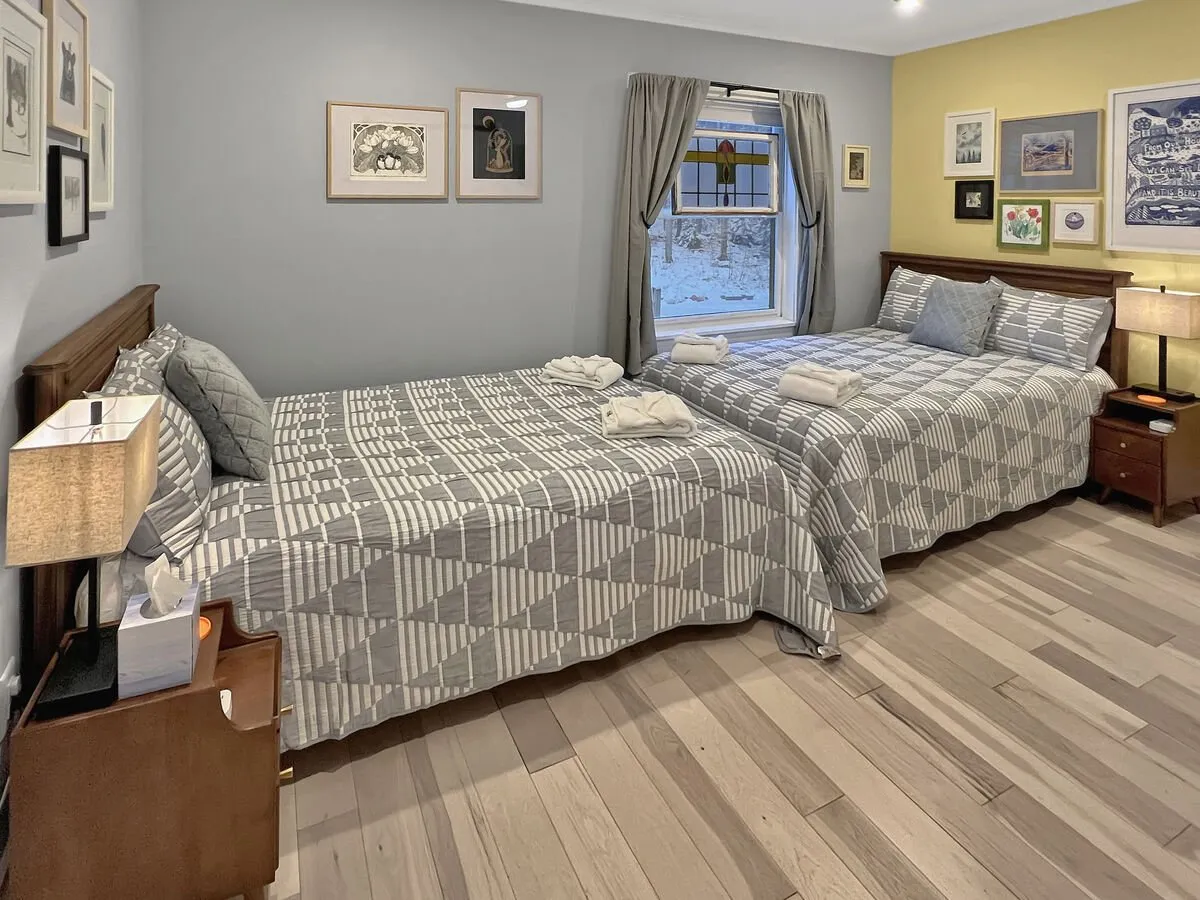 A warmly lit bedroom showcasing two full beds with patterned quilts, decorative wall art, and a window letting in natural light. The space exudes a cozy, welcoming mood, ideal for families or friends sharing the room.