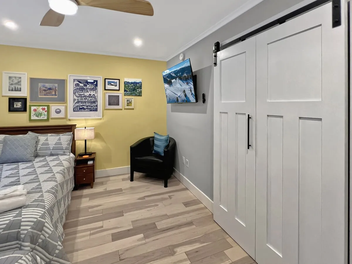 A stylish bedroom with a patterned bedspread, a gallery wall filled with art, a modern sliding barn door, and a TV. The space feels inviting and functional, offering a spot to relax and recharge. Subtle hints of the outdoors are provided through decor and the barn door, hinting at the region's rustic charm.