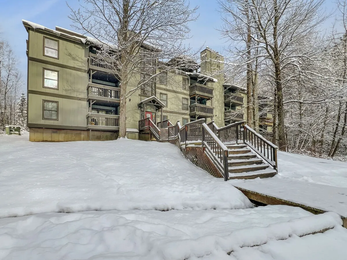 The exterior of the condo complex during winter, with a peaceful snow-covered ground, a sturdy wooden staircase, and a green facade nestled among bare trees. The setting evokes a serene retreat, ideal for returning after skiing or winter adventures.
