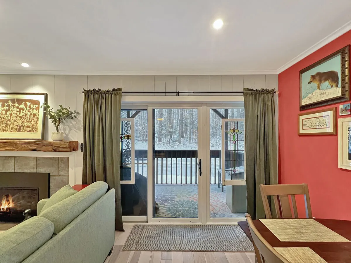 This image captures a welcoming interior living area featuring a large sliding glass door that opens onto a balcony or patio. The cozy fireplace, wooden dining table, and green curtains create an inviting atmosphere, while the snow-capped view outside hints at the tranquil mountain and woodland setting described in the section.