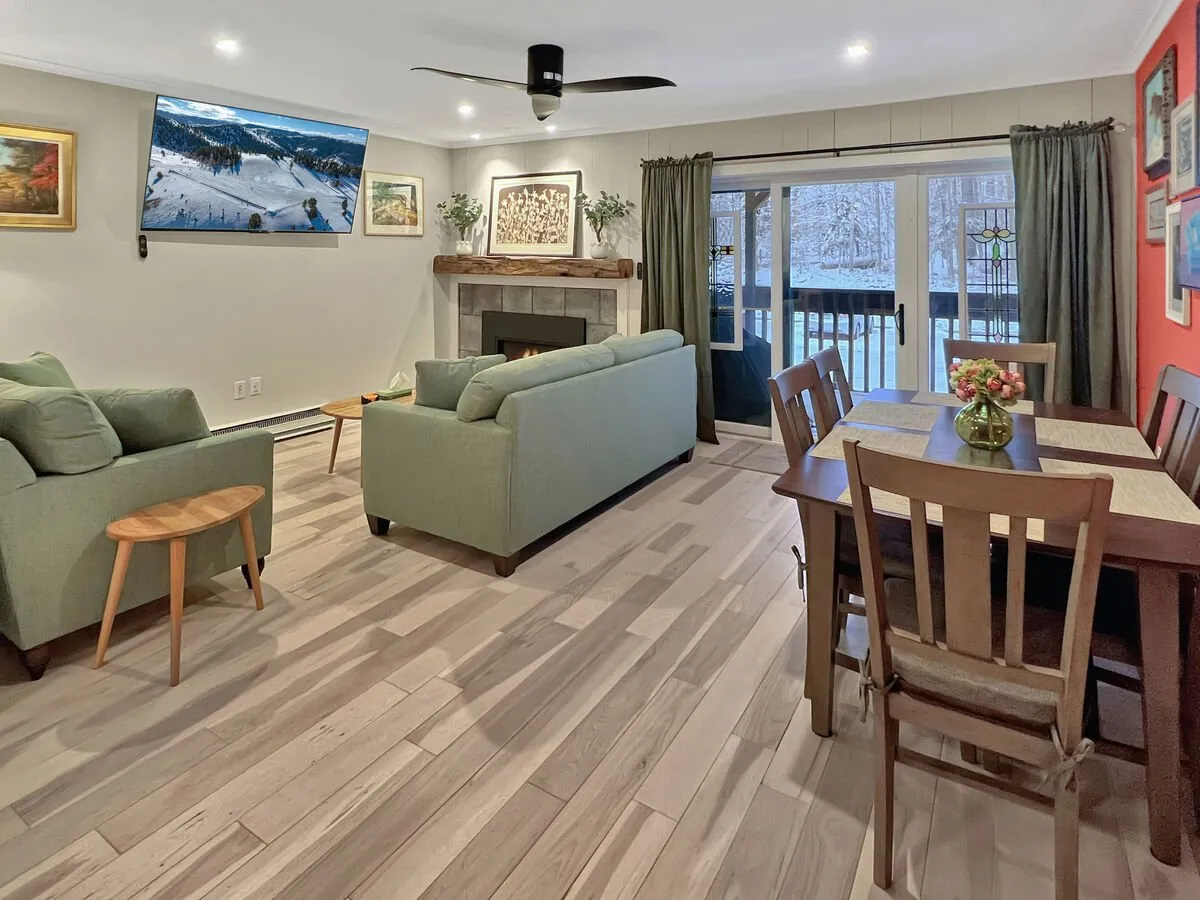 The image shows a spacious, warmly-lit living and dining area in the condo, with comfortable sofas arranged around a crackling fireplace, a family-style dining table, tasteful artwork, and large windows that let in natural light and glimpses of Vermont’s greenery. The scene evokes comfort and togetherness, perfect for cozy gatherings or relaxing evenings after outdoor adventures.