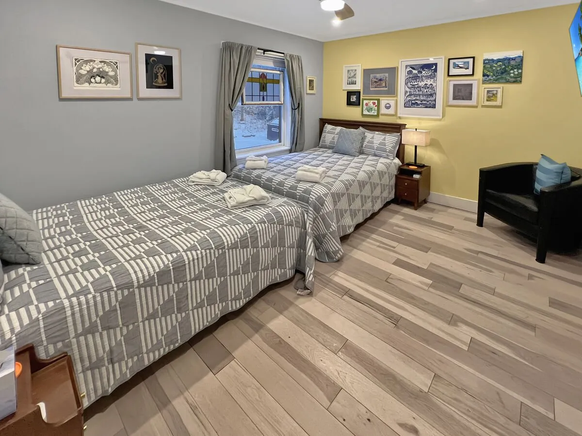 This image features a welcoming room with two twin beds adorned in patterned bedding, a cozy black armchair, and a large window streaming in natural light. The window, though the exterior view is limited, suggests an inviting connection to the outdoors, while the bright, airy atmosphere hints at restful evenings after a day spent exploring.