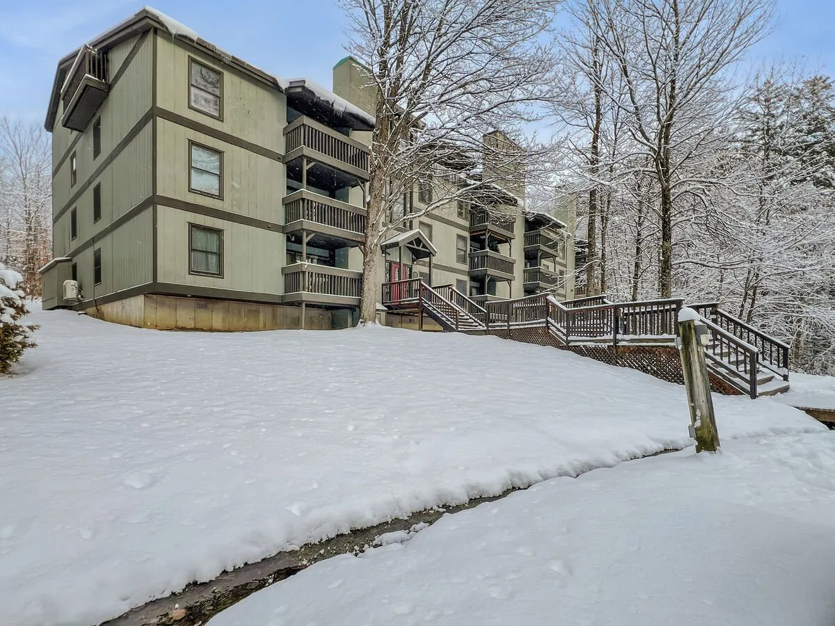 An exterior view of the property, showcasing snow-covered ground, multiple balconies, wooden stairs, and surrounding trees. The image emphasizes the peaceful natural environment and highlights private outdoor spaces designed for enjoying scenic serenity.