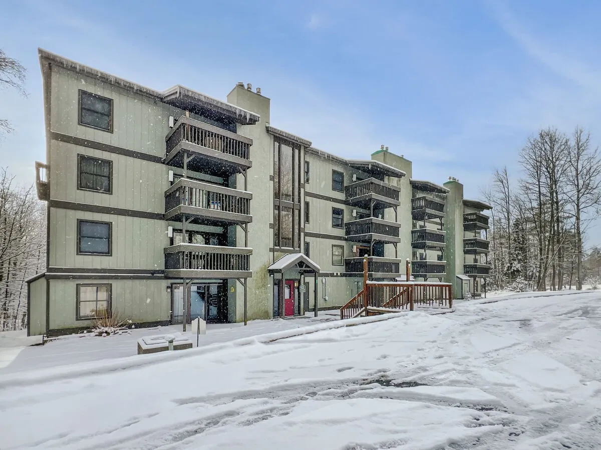 A charming exterior angle highlighting the red-doored entrance, snow-coated balconies, and mature trees. This inviting scene conveys the sense of community and warmth, while illustrating the opportunities for relaxing or socializing on the balcony amidst tranquil surroundings.