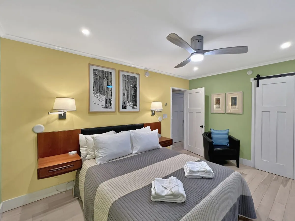 A cozy master bedroom featuring a queen bed with decorative linens, a contemporary ceiling fan, and nature-inspired wall art. A side table with a lamp sits next to the bed, and soft daylight streams softly into the room, creating a tranquil atmosphere.